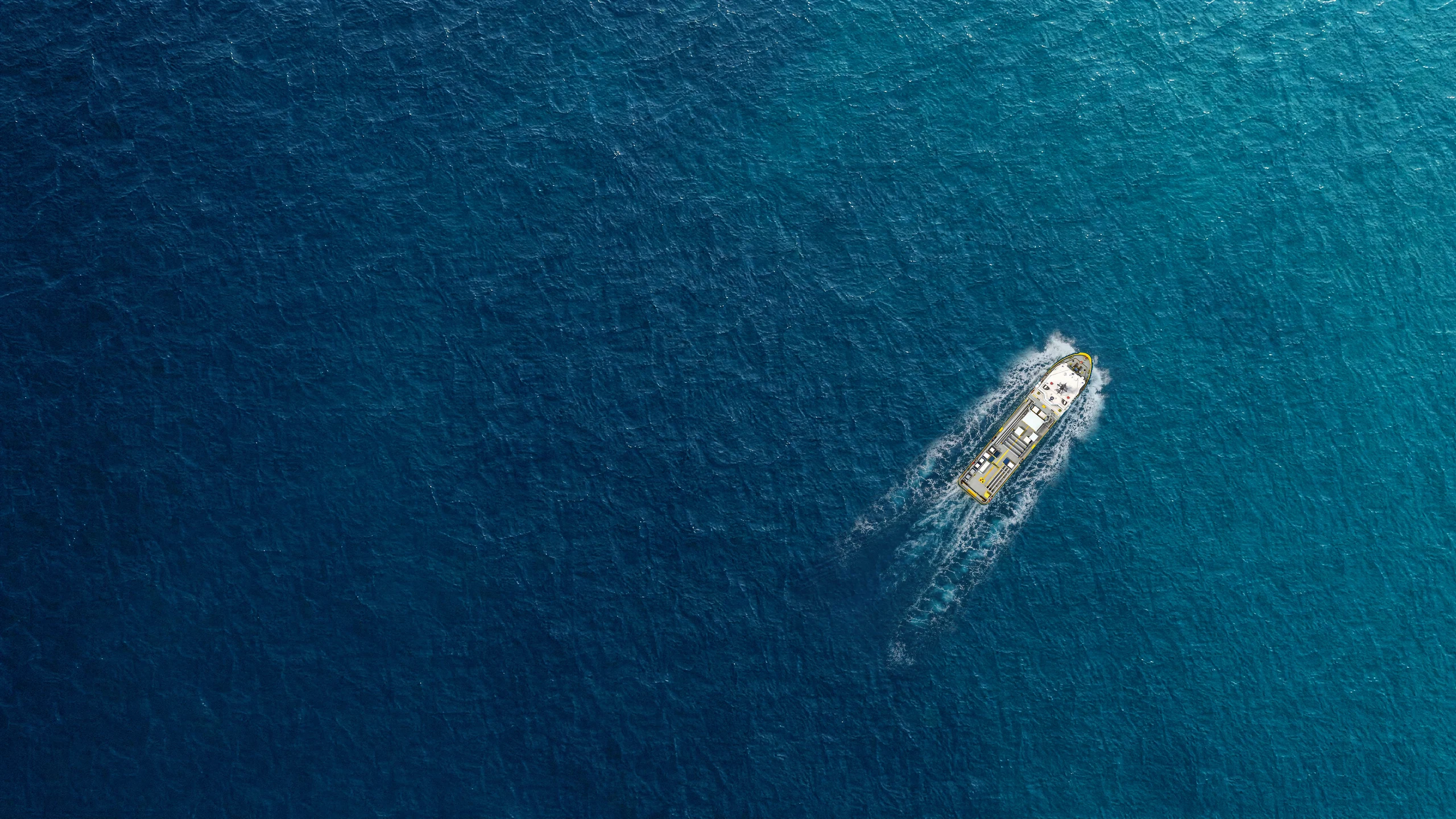 Overhead view of a navy and yellow offshore vessel underway, with bow foam and a wake trailing through blue ocean water
